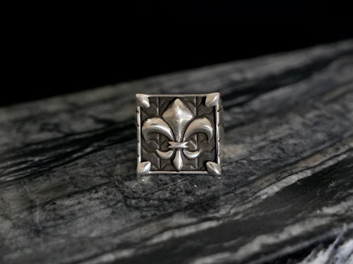 A square silver-toned signet ring featuring a raised fleur-de-lis motif, photographed on a dark marbled surface against a black background.