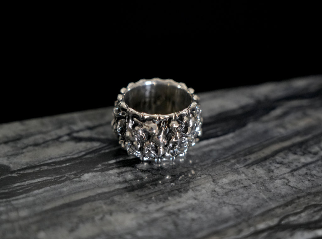 A wide silver ring with an intricate, textured Veles animal skulls design, resting on a dark, polished stone surface against a solid black background.