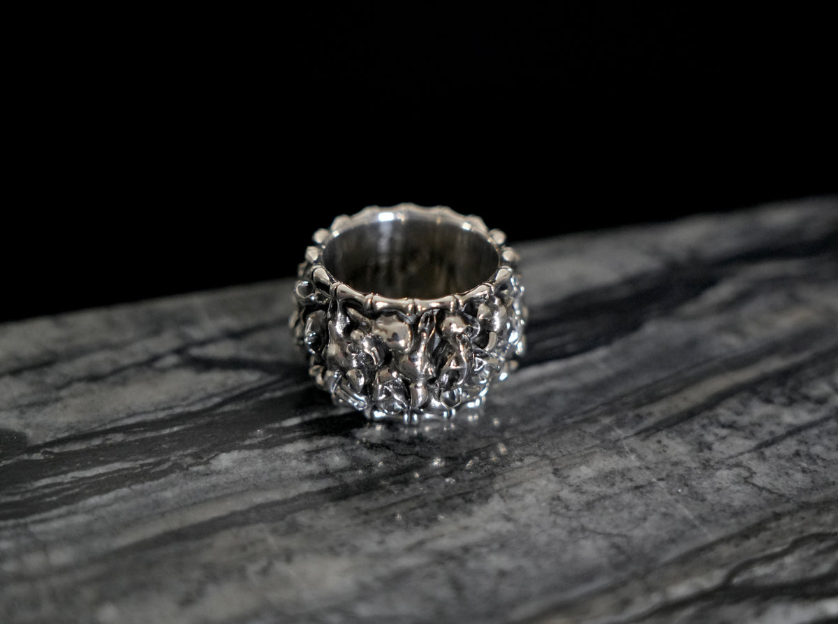 A wide silver ring with an intricate, textured Veles animal skulls design, resting on a dark, polished stone surface against a solid black background.